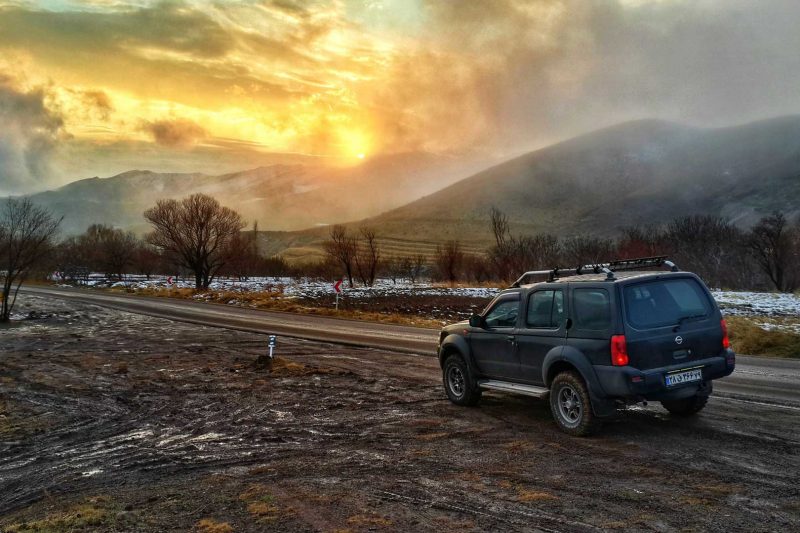 A Nissan Xterra parked on the side of a dirt road as the sun rises over foggy mountains.