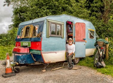 A man sits in an old blue and white pop up camper with a for sale sign attached to the front.