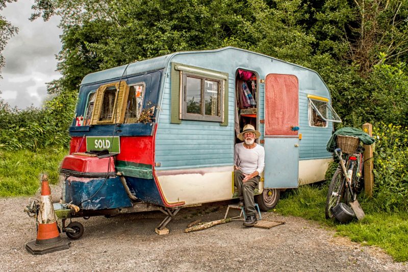 A man sits in an old blue and white pop up camper with a for sale sign attached to the front.