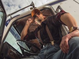 A truck driver opens the cabin door and looks out at a truck stop with his sunglasses on.