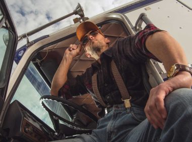A truck driver opens the cabin door and looks out at a truck stop with his sunglasses on.
