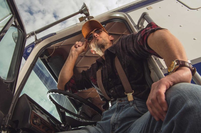 A truck driver opens the cabin door and looks out at a truck stop with his sunglasses on.