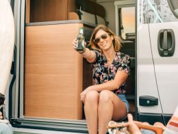 A woman drinks a beer while in her camper van surrounded by friends.
