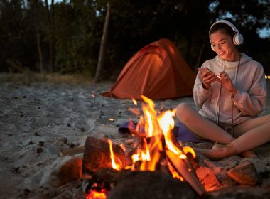 A woman sits by a campfire with her headphones on listening to a podcast.