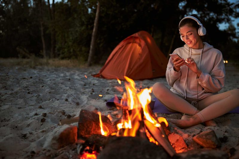 A woman sits by a campfire with her headphones on listening to a podcast.