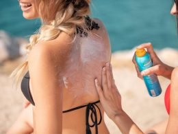 A friend applies sunscreen to her friends back on the beach.