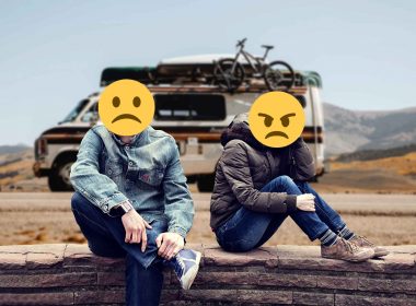 A couple in an argument sitting on a stone wall in front of an RV with sad and angry emojis over their faces.