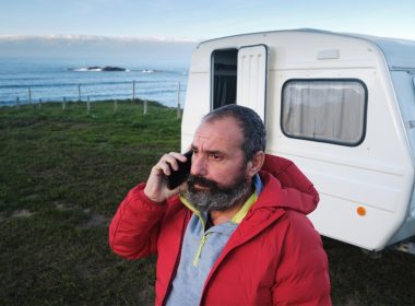 A bearded man makes a concerned phone call in front of his travel trailer.