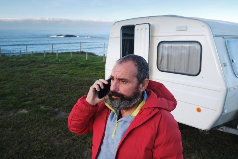 A bearded man makes a concerned phone call in front of his travel trailer.