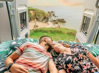 Young couple kissing in the bed of their campervan with a view of the ocean behind them.