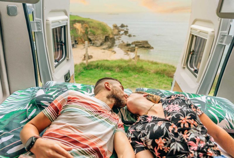 Young couple kissing in the bed of their campervan with a view of the ocean behind them.