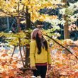 A woman in a bright yellow jacket stands in a forest full of leaves changing colors in fall.