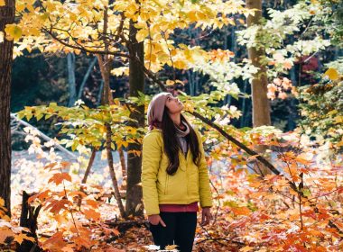 A woman in a bright yellow jacket stands in a forest full of leaves changing colors in fall.