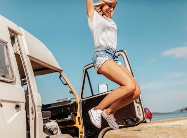 A woman jumping up in the air with her hands up in excitement next to her camper van on the beach.