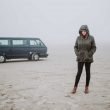 A woman with her hood up stands outside of her conversion van and looks sadly down at the ground, surrounded by fog.