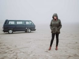 A woman with her hood up stands outside of her conversion van and looks sadly down at the ground, surrounded by fog.