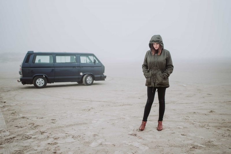 A woman with her hood up stands outside of her conversion van and looks sadly down at the ground, surrounded by fog.