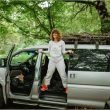 A woman stands in the side door of her van smiling and shrugging while parked in a wooded park.