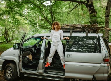 A woman stands in the side door of her van smiling and shrugging while parked in a wooded park.
