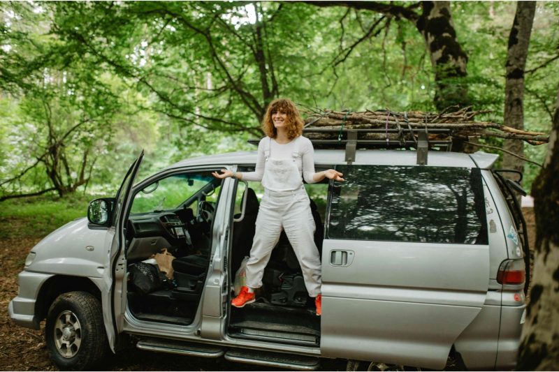 A woman stands in the side door of her van smiling and shrugging while parked in a wooded park.