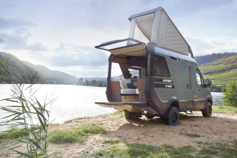 Hymer's concept car VisionVenture camper van parked next to a beautiful lake on a summer day, with the popup roof and back patio doors open.