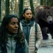 Two women hikers look up fearfully in low lit woods as a large brown bear approaches from behind them in the woods.