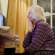 An elderly woman holds her hand over her mouth, troubled, as she looks at her desktop computer screen.
