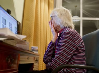 An elderly woman holds her hand over her mouth, troubled, as she looks at her desktop computer screen.