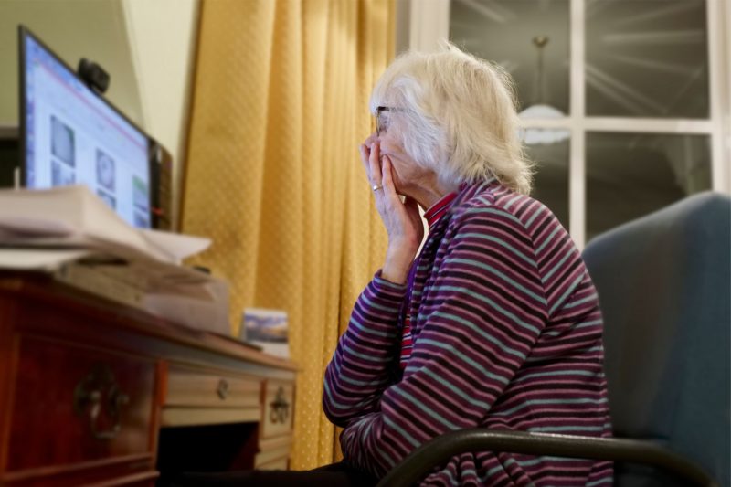 An elderly woman holds her hand over her mouth, troubled, as she looks at her desktop computer screen.