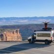 A man stands on top of his camper van RV with his arms stretched out to show the expansive size of the mountain view behind him.