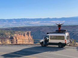 A man stands on top of his camper van RV with his arms stretched out to show the expansive size of the mountain view behind him.