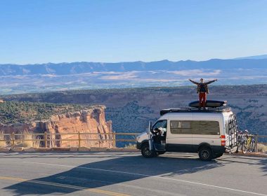 A man stands on top of his camper van RV with his arms stretched out to show the expansive size of the mountain view behind him.
