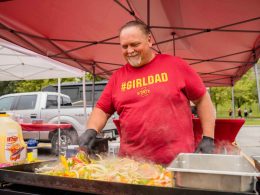 A dad wearing a #girldad shirt grills up food on a flat top in a parking lock during a tailgate party.