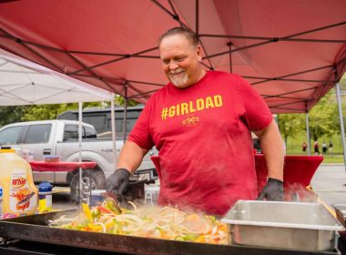 A dad wearing a #girldad shirt grills up food on a flat top in a parking lock during a tailgate party.