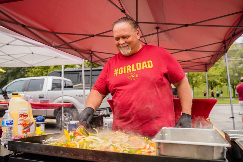 A dad wearing a #girldad shirt grills up food on a flat top in a parking lock during a tailgate party.