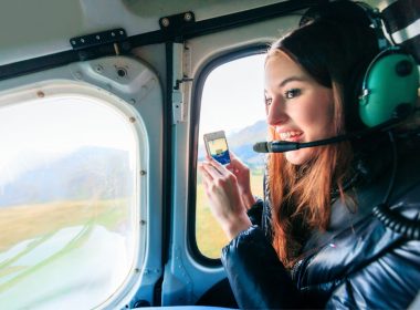 A tourist wearing a headset takes a picture on her phone as she looks out the window of a helicopter.
