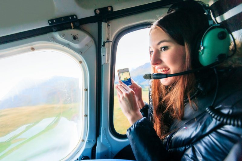 A tourist wearing a headset takes a picture on her phone as she looks out the window of a helicopter.