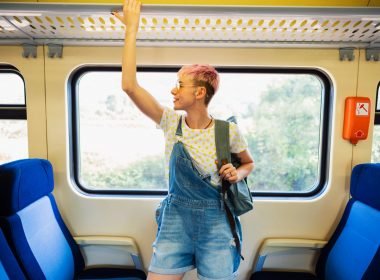 A person holds onto the luggage rack on a train as they smile in anticipation of their travels.