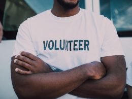 A man stands with his arms crossed wearing a white volunteer shirt.