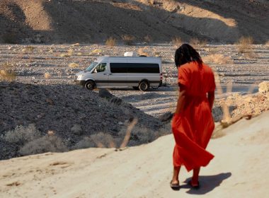 A woman looks down at her feet and her van is parked in the distance in the desert.