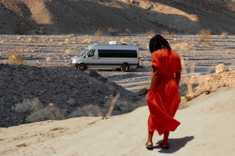 A woman looks down at her feet and her van is parked in the distance in the desert.