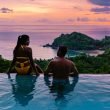 A couple on vacation relaxing in a pool overlooking the ocean at sunset.