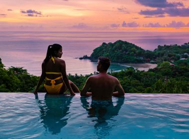 A couple on vacation relaxing in a pool overlooking the ocean at sunset.