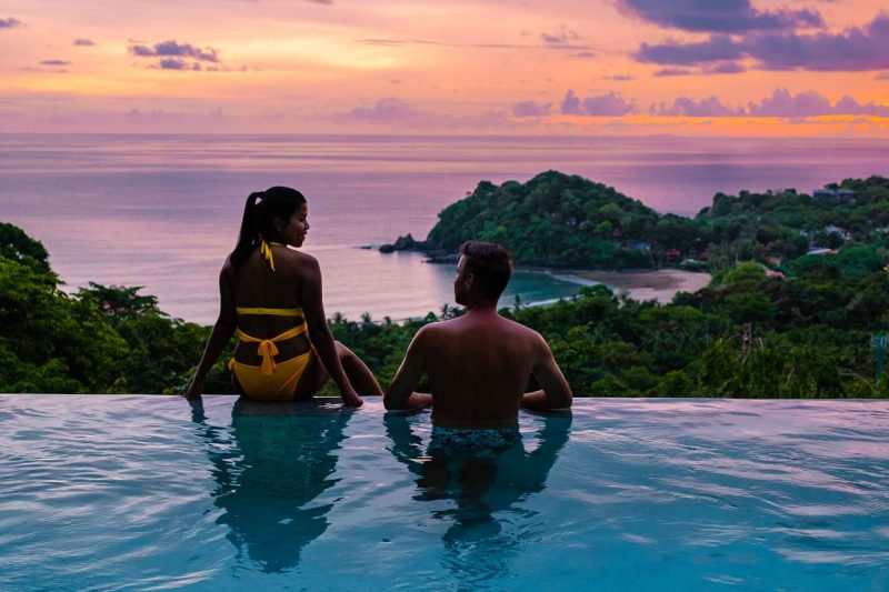 A couple on vacation relaxing in a pool overlooking the ocean at sunset.