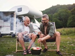 An adult man and his father share a beer while they cook food over the father at an RV Park.