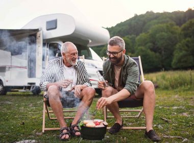 An adult man and his father share a beer while they cook food over the father at an RV Park.