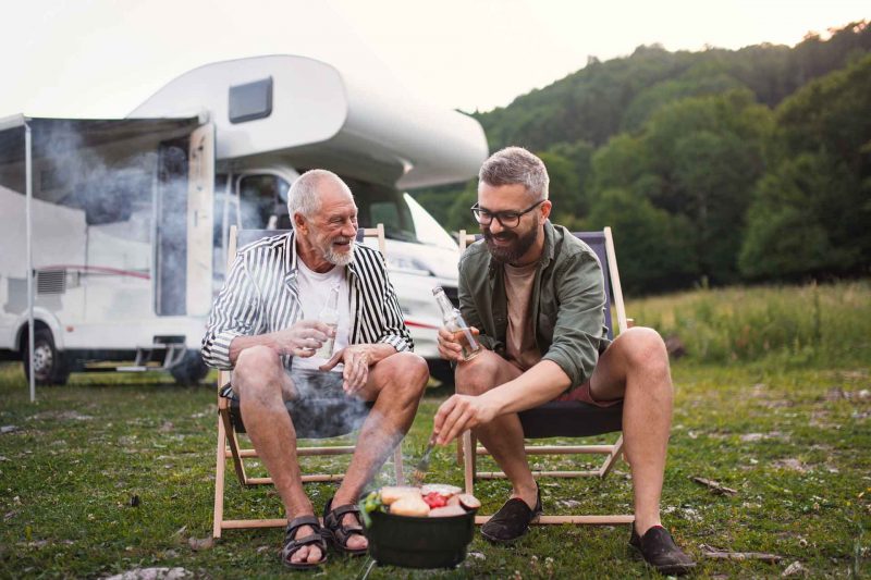 An adult man and his father share a beer while they cook food over the father at an RV Park.