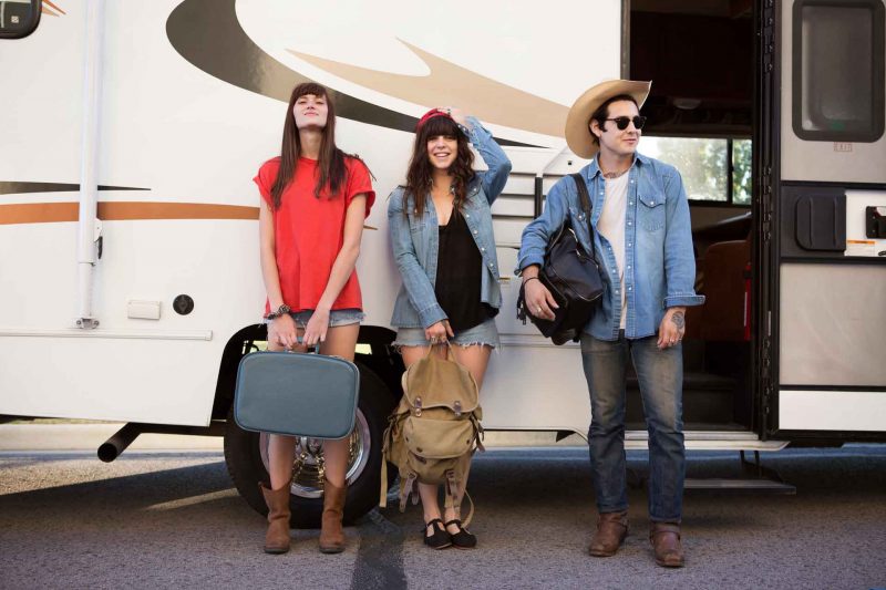Three friends standing with their luggage in front of an RV, ready to go on a cross country trip.