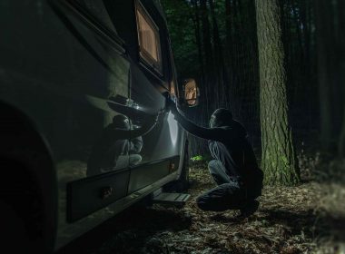 A burglar attempts to pick the lock on a motorhome door parked in the woods at night.