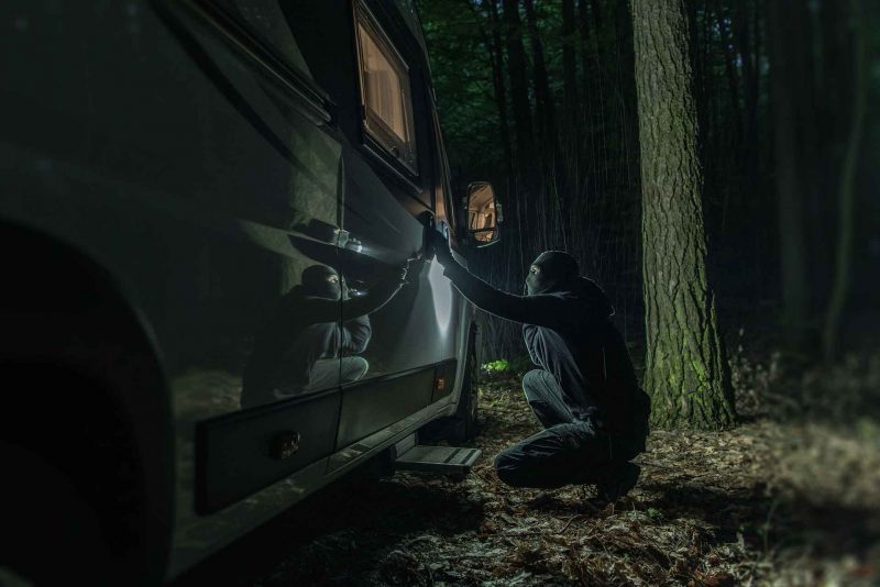 A burglar attempts to pick the lock on a motorhome door parked in the woods at night.